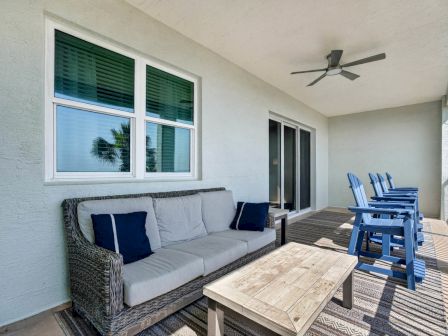 A bright patio with a gray outdoor sofa, blue chairs, a wooden table, ceiling fan, and large windows.