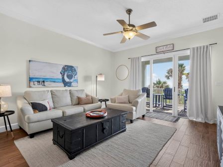 A bright living room with white sofas, a dark coffee table, a beach-dog wall painting, a ceiling fan, and glass doors to a balcony.