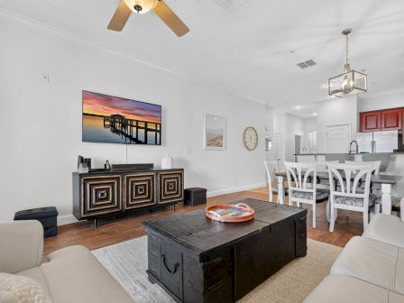 Bright living-dining room with a TV, wall art, dark console, coffee table, white sofas, dining set, and hanging chandelier.