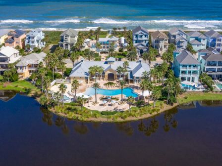 Aerial view of a coastal neighborhood with colorful houses, a central pool area, palm trees, and a waterway surrounding the community near the ocean.