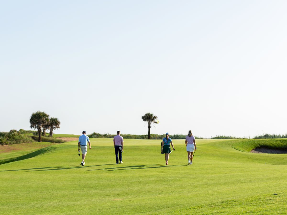 Four people walk side by side on a sunny golf course, green fairways, a few trees in the distance, and a clear blue sky.