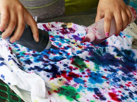 People tie-dyeing a white T-shirt with colorful dye splashes, using bottles to squirt and blend bright blues, greens, reds, and purples.