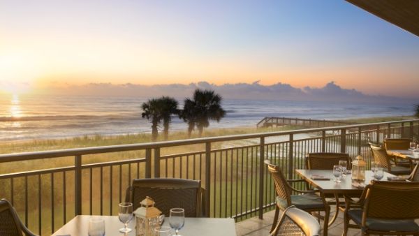A scenic beachfront dining area with set tables, overlooking the ocean and palm trees at sunrise or sunset.