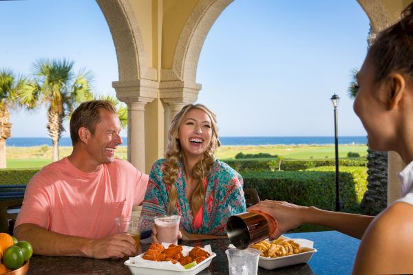 A group of people enjoys a meal and drinks outdoors near the ocean, with palm trees and a clear sky in the background.