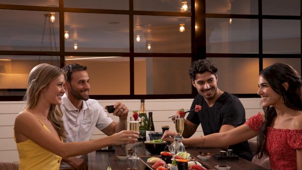 Four people enjoying sushi and drinks at a restaurant, smiling and engaging in conversation in a warmly lit setting.
