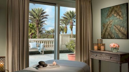 A cozy room with a massage table, candles, and art on the wall. Large windows offer a view of palm trees outside, creating a relaxing ambiance.