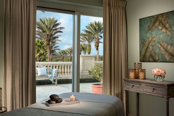 A cozy room with a massage table, candles, and art on the wall. Large windows offer a view of palm trees outside, creating a relaxing ambiance.