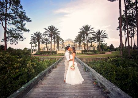 A couple is embracing on a wooden bridge lined with palm trees, with a grand building in the background under a partly cloudy sky.