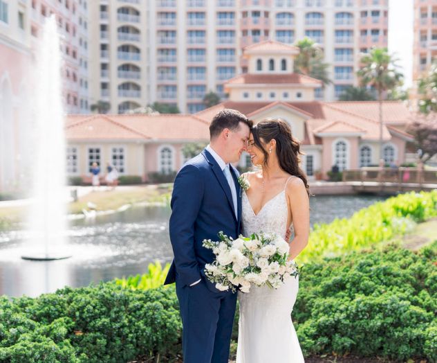 A bride in a white gown and a groom in a navy suit share a kiss beside a garden fountain, holding a bouquet, with a resort backdrop.