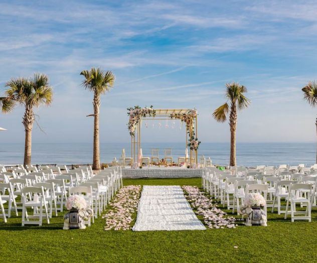 A beachfront wedding setup with a white aisle, chairs, palm trees, and an arbor against the ocean and blue sky backdrop.