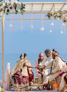 A wedding ceremony takes place outdoors under a floral arch by the sea, with traditional attire worn by participants.