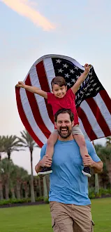 A smiling dad lifts his child outdoors with an American flag, celebrating at sunset.