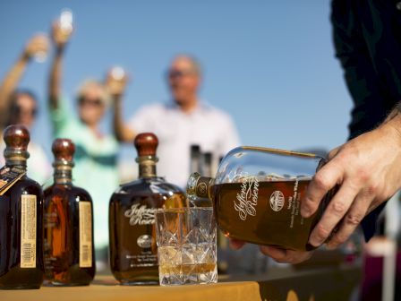 A man pours amber liquor from a bottle into a cut-glass tumbler on a table, with other bottles and blurred people in the background.