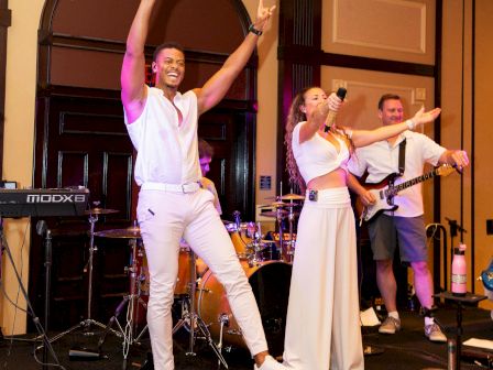 A singer and a guitarist band perform on a hotel stage, cheering with raised arms and microphones under warm lights.
