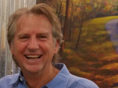 A smiling older man in a blue shirt, indoors beside a colorful autumn painting.