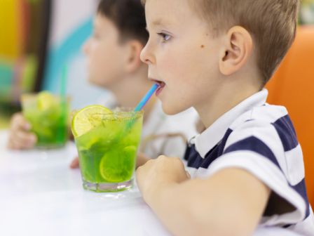 A boy sips a bright green drink through a blue straw at a table while someone sits behind him.