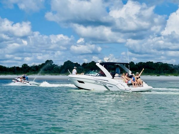 A group of friends rides a luxury speedboat on a river while another boat follows under a bright blue, cloudy sky.