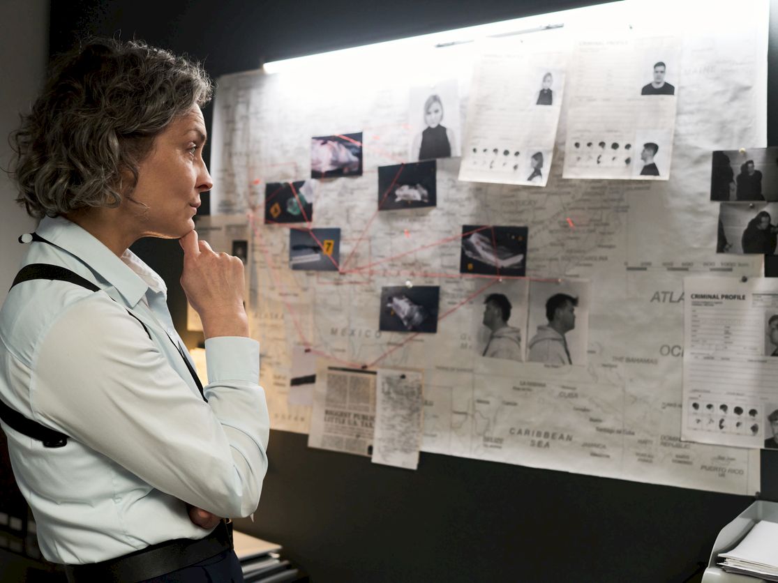 A woman in an office studies a wall covered with criminal photos, mugshots, and maps, deep in thought.