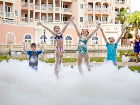 Kids in swimsuits laugh and jump through foam at a sunny resort pool, cheering with a big splash in front of balconies.