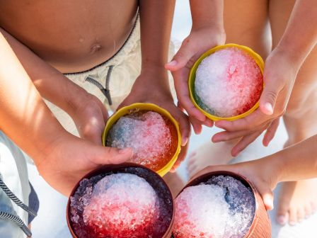 Friends hold bowls of colorful shaved ice in the sun, cheering together around the beach.