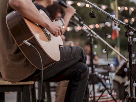 A guitarist strums an acoustic on a cozy outdoor stage, mic stands, string lights, and a small audience in the background.
