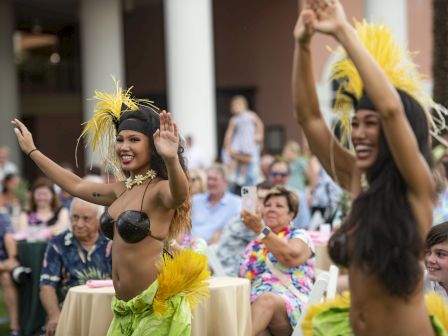 A performer in a yellow-dressed costume waves pom-poms at an outdoor celebration while cheering spectators watch.