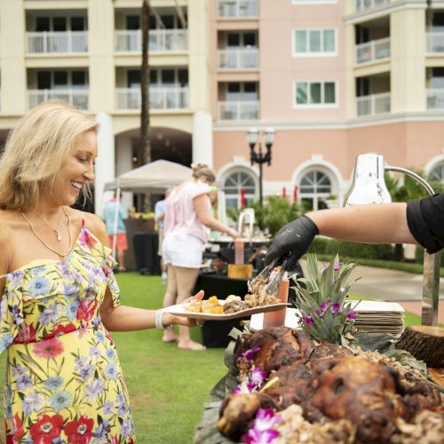 An outdoor reception shows a server in gloves pouring food from a pitcher to a guest at a table, with other attendees dining nearby.