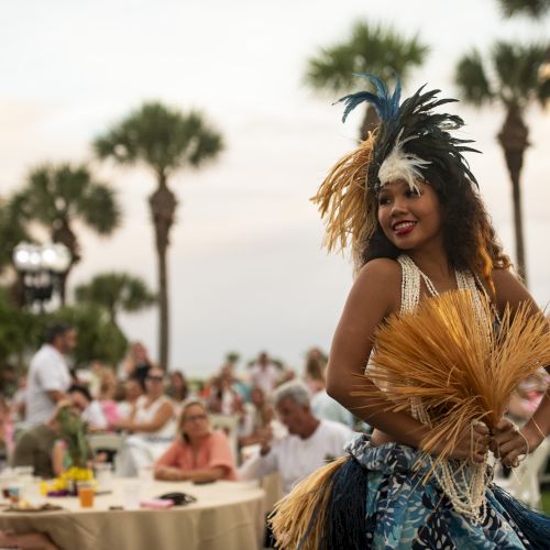A performer in a colorful feathered headdress and grass skirt dances at an outdoor event while a crowd watches.