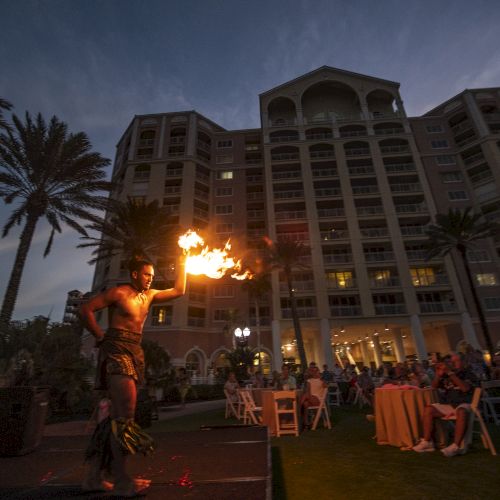 A fire-dancer woman spins flames with a torch in a courtyard before a lit hotel building at dusk.