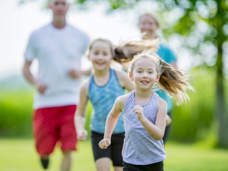 A little girl runs smiling outdoors in a park while other family members follow in the background.