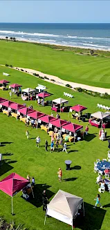 An outdoor event on a golf course by the sea features numerous tents, tables, and people gathered on the grassy area.