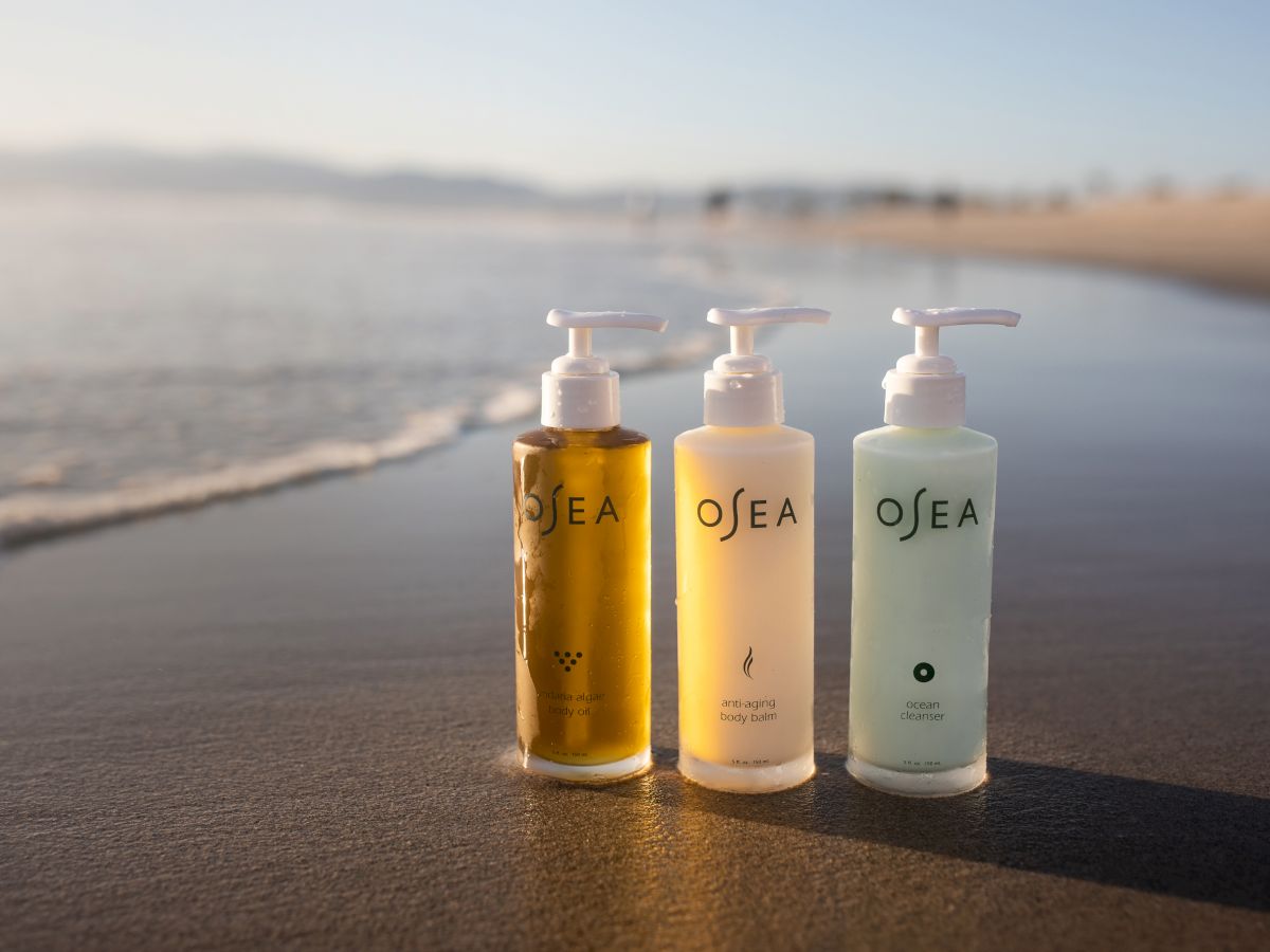 Three pump-top OSEA skincare bottles&mdash;body oil, body balm, and ocean cleanser&mdash;lined up on a sunlit beach.