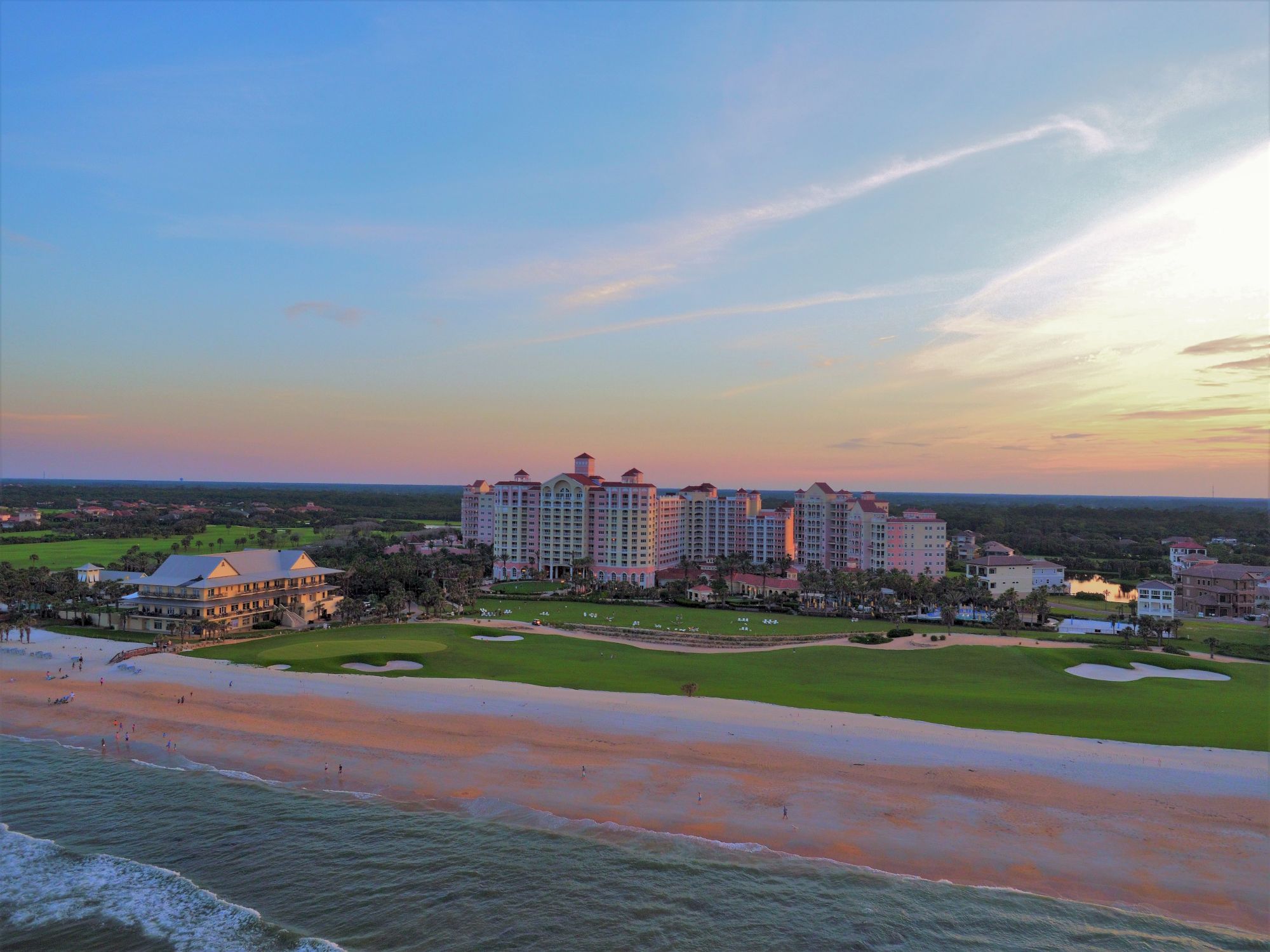 A coastal landscape with a beach, multiple buildings, and a golf course at sunset under a colorful sky.