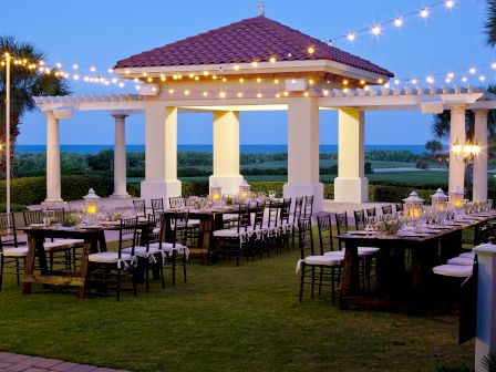 An outdoor dining area with tables and chairs, set under string lights and a pavilion, against a scenic backdrop of trees and ocean.