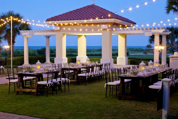 An outdoor dining area with tables and chairs, set under string lights and a pavilion, against a scenic backdrop of trees and ocean.