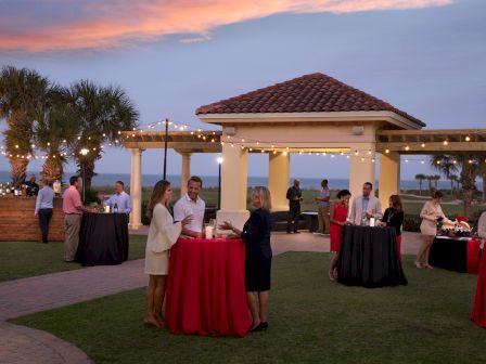 People mingle at an outdoor evening cocktail party by a gazebo, servers and tall round tables with red and black linens under string lights.