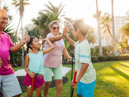 A family enjoying mini-golf with smiles and high-fives under the sun, surrounded by palm trees and greenery.