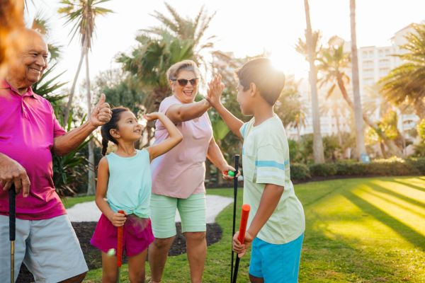 A family enjoying mini-golf with smiles and high-fives under the sun, surrounded by palm trees and greenery.