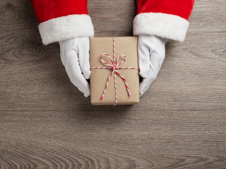 Santa-clad hands present a wrapped gift box with red-and-white striped ribbon on a wooden floor.