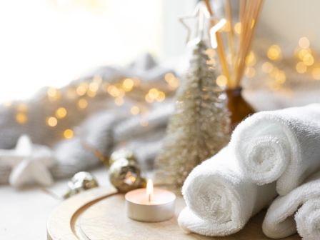 A spa-like winter scene with rolled white towels beside a lit candle, faux pine tree, and golden holiday bokeh lights.