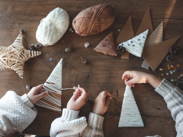 Kids&rsquo; hands decorate a wooden table with yarn crafts&mdash;little Christmas trees and ornaments made from twine and pinecones.