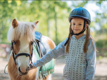 A young child in a helmet holds the reins while riding a white horse outdoors, looking calm and confident.