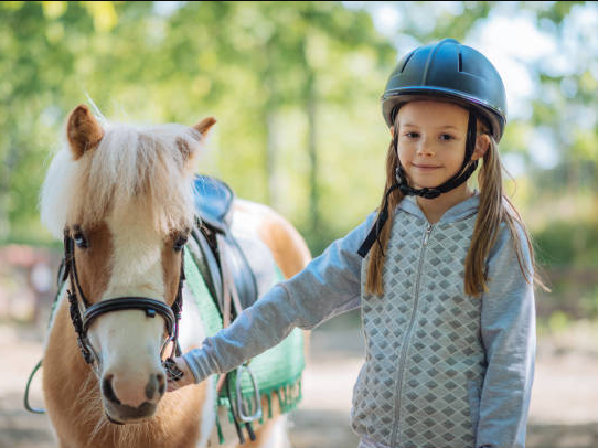 A young child in a helmet holds the reins while riding a white horse outdoors, looking calm and confident.