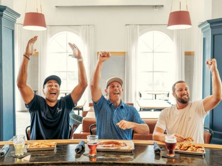 Three men at a table in a bright room cheer with fists raised, celebrating together while drinks and food sit in front of them.