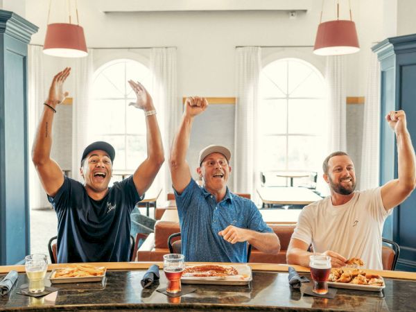 Three men at a table in a bright room cheer with fists raised, celebrating together while drinks and food sit in front of them.