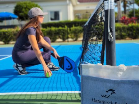 A woman sets up a pickleball net on a poolside court under a blue, cloudy sky.