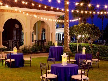 An outdoor setting with round tables covered in purple cloth, surrounded by chairs, lanterns on tables, and string lights overhead for ambiance.