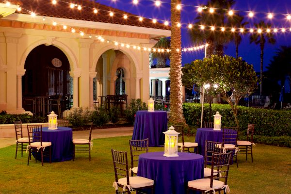 An outdoor setting with round tables covered in purple cloth, surrounded by chairs, lanterns on tables, and string lights overhead for ambiance.
