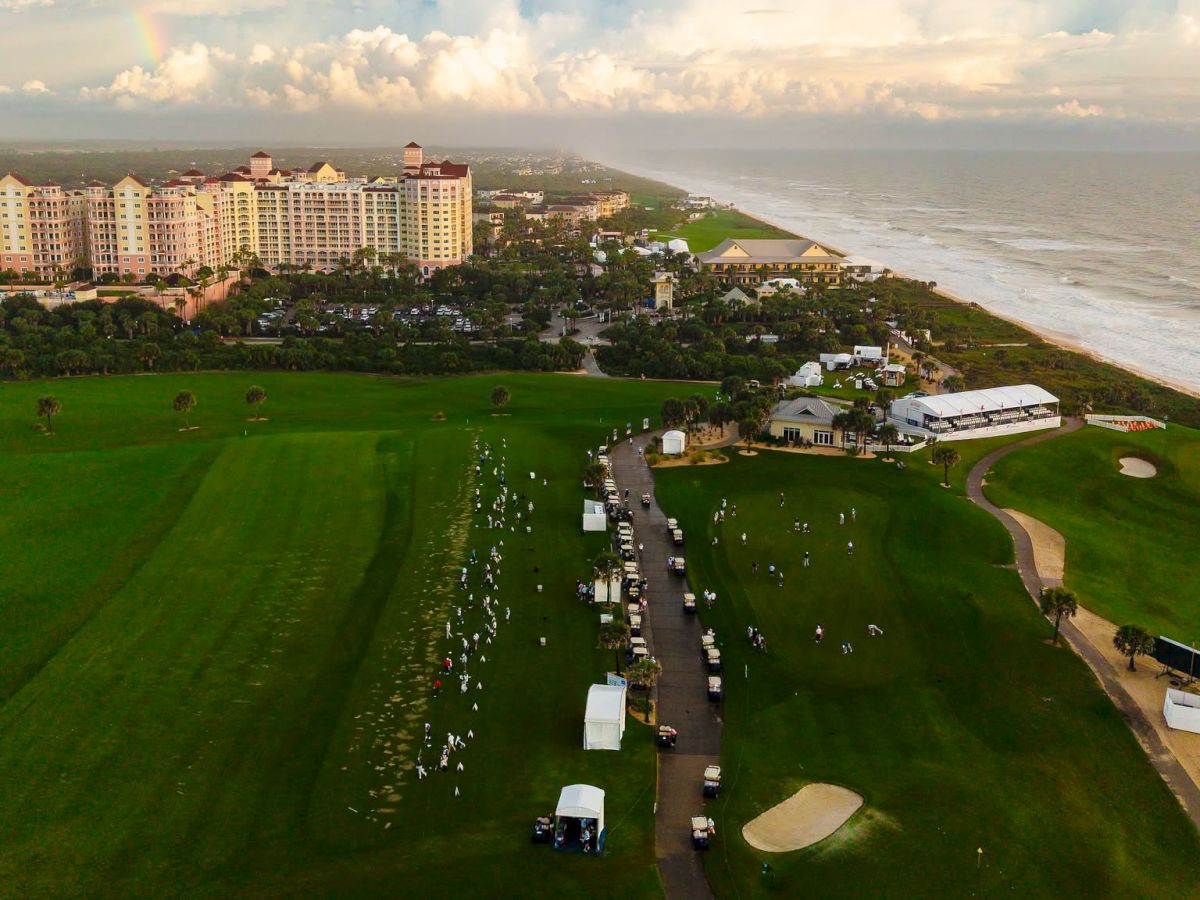 Aerial view of a coastal resort with lush lawns, event tents, people gathering, and apartment buildings along the shoreline under clouds.