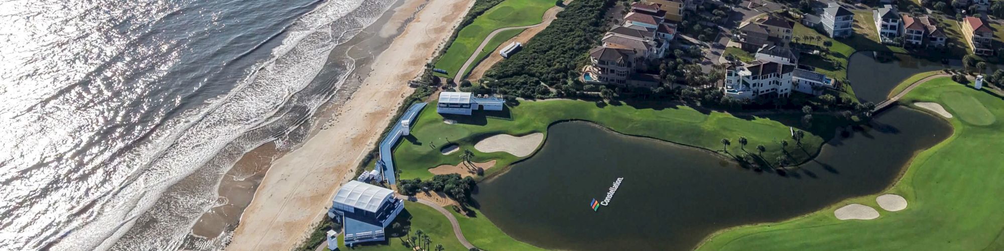An aerial view of a seaside golf course with manicured greens, ponds, and beachfront waves beside coastal buildings and homes.